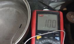Czerwony miernik marki UNI-T UT70A pokazujący temperaturę 100°C obok metalowego garnka z wodą i sondą termopary w zanurzeniu.