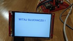 LCD module on a red PCB showing “WITAJ SŁUCHACZU!” in blue on a light background, on a wooden table.