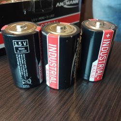 Trzy baterie alkaliczne Ansmann Mono D-LR20 1.5V ustawione w rzędzie na drewnianym stole.