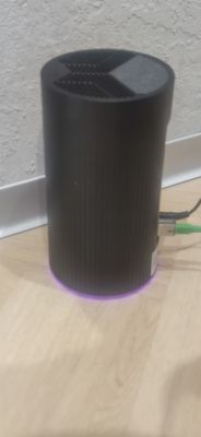 Cylindryczny router Playbox z fioletowym światełkiem na podstawie, umieszczony na podłodze przy ścianie.