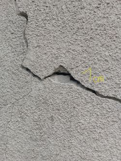 Pionowe i poziome pęknięcia na powierzchni betonowej ściany z widoczną szczeliną o szerokości około 1 cm.
