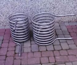 Dwie wężownice ze stali nierdzewnej stojące na kostce brukowej przy ścianie.