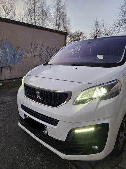 Biały Peugeot Expert z włączonymi światłami przednimi na tle ściany z graffiti.