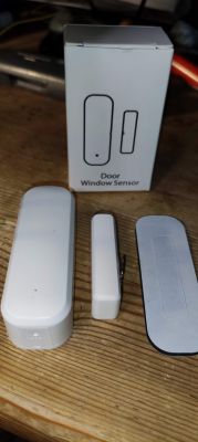 SZKOSTON Door/Window Sensor with Packaging SZKOSTON door/window sensor set next to packaging.