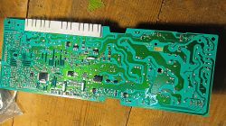 Moduł elektroniczny pralki z widocznymi ścieżkami PCB, leżący na drewnianym blacie, bez widocznych uszkodzeń termicznych.