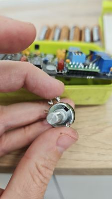 Potencjometr z gładkim wałkiem trzymany w dłoni Palce trzymają metalowy potencjometr z gładkim wałkiem; w tle płytka elektroniczna w zielonej obudowie.