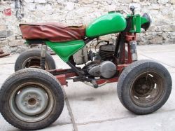 Domowej roboty trójkołowiec z silnikiem motocyklowym i zielonym zbiornikiem paliwa, pośród kamiennego podwórka.