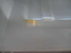 Pęknięcie w plastiku pod szufladą lodówki z żółtą taśmą na uszkodzeniu.