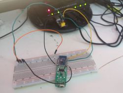 Breadboard with a small circuit board and jumper wires on a desk; device with LED lights in the background.