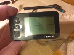Wyświetlacz LCD roweru elektrycznego z przyciskami po lewej stronie (+, M, -) i logo Indiana na dole.