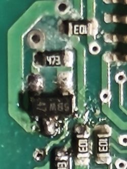 Zbliżenie na płytkę PCB z elementami SMD; jeden układ wygląda na przypalony, widać ścieżki i luty.