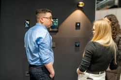 Two people talk in front of a black wall with control panels and a wall light