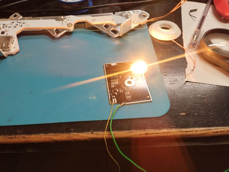 Płytka PCB z diodą LED emitującą jasne światło, podłączona przewodami, leżąca na niebieskiej podstawce obok elementów lampy samochodowej.