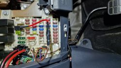 Widok na skrzynkę bezpieczników w samochodzie z dodatkowym czerwonym kablem podłączonym do jednego z bezpieczników.