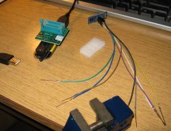 Adapter programatora podłączony przez USB, zestaw przewodów z cienkimi szpilkami oraz kawałek białej pianki na drewnianym biurku.