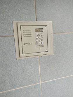 Panel domofonowy Cyfral zamontowany na ścianie z szarej glazury.