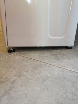 Dolna część pralki pokazująca nóżki ustawione na płytkach ceramicznych.