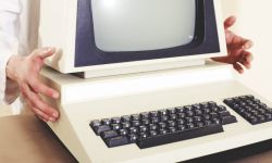 Ręce trzymają beżowy komputer z monitorem CRT na obudowie i czarną klawiaturą