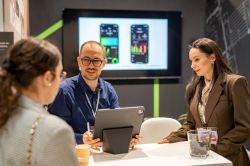 Three people at a table discuss a tablet; a screen behind shows a mobile app interface.