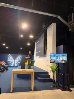 Speaker at a podium on a conference stage with a large screen and audience on the left