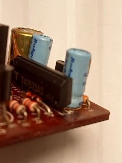 Zbliżenie na płytkę drukowaną z widocznymi kondensatorami elektrolitycznymi, rezystorami i układem scalonym TA7523AS; na jednym kondensatorze brązowa, wyciekająca substancja.