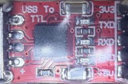 Close-up of a USB to TTL converter module on a red PCB with labeled signal connections and components.