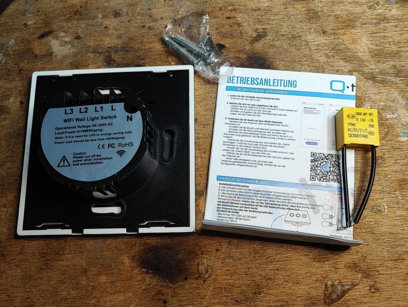 QTouch WiFi Wall Switch Set with Accessories Set containing a WiFi wall switch, capacitor, and instruction manual on a wooden table.