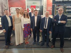 Grupa ośmiu osób na stoisku targowym firmy RST z ekspozycją rozdzielnic i urządzeń ochronnych.