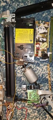 Rozebrane podzespoły drukarki i elektronika na kocu, m.in. moduł z ostrzeżeniem i silnik.