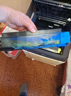 Osoba trzymająca pojemnik na toner z widocznymi śladami rozlanego tonera w tle z otwartą drukarką Kyocera.