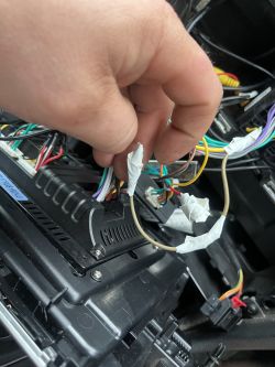 Ręka trzymająca przewody elektryczne w samochodzie, widoczne są różnokolorowe kable i plastikowe osłony.