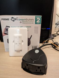 Prime WiFi Smart Outdoor Outlets with Remote Control Black Prime WiFi Smart Outdoor Outlets adapter with remote control against product box.