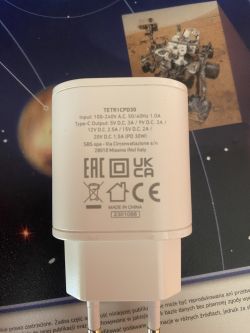 Biały adapter ładowarki z oznaczeniami technicznymi na tle zdjęcia łazika.