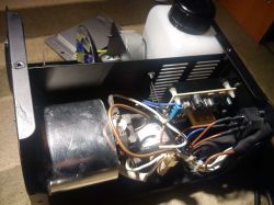 Wnętrze urządzenia z płytką elektroniki, okablowaniem, metalową komorą i białym zbiornikiem z czarną nakrętką