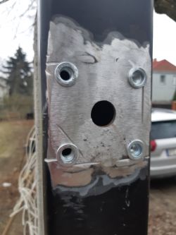 Metalowy słupek z otworami na śruby zamontowany przy bramie.