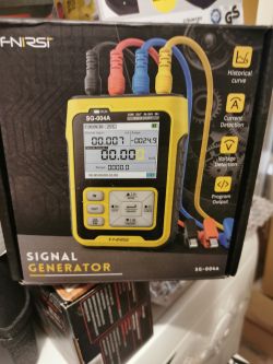 Generator sygnału FNIRSI SG-004A w żółtej obudowie, na opakowaniu widać przewody i wyświetlacz LCD z pomiarami.