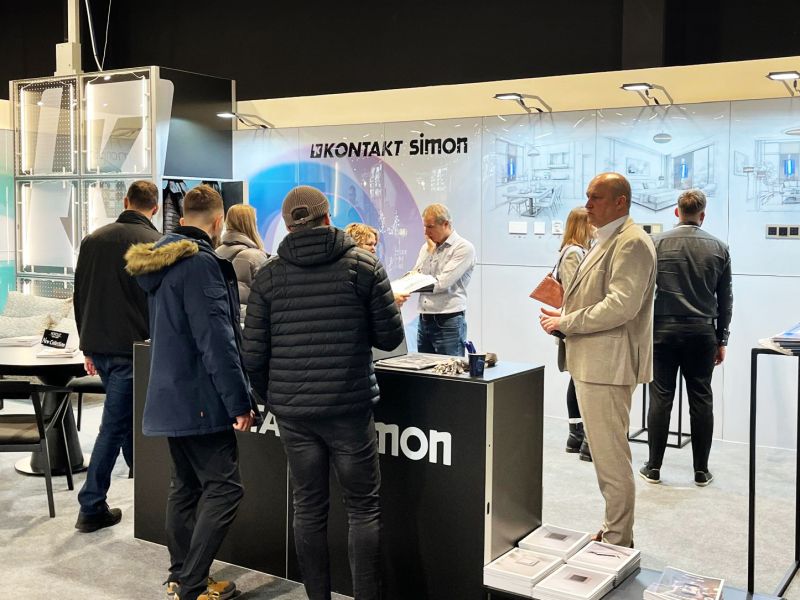 Trade show booth “Kontakt Simon” with visitors, a counter, display panels, and interior renderings on the back wall.