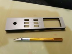 Metalowy panel z wycięciami i okrągłym otworem, obok złoty nożyk modelarski na blacie