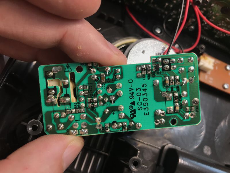 Bottom of a green PCB held between fingers, with solder joints and markings “94V-0 SC-03 E550345”.