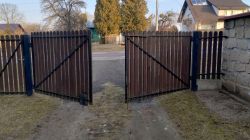Otwarta brama skrzydłowa wykonana z drewna, z metalowymi wzmocnieniami, prowadząca na zewnątrz na drogę.