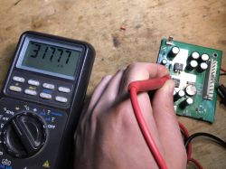 Ręka trzymająca sondę multimetru, który mierzy częstotliwość 317,77 kHz na płytce elektronicznej z kondensatorami.