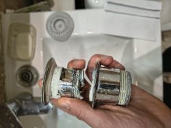 Dłoń trzyma pęknięty metalowy element odpływu z gwintem, rozdzielony na dwie części nad umywalką