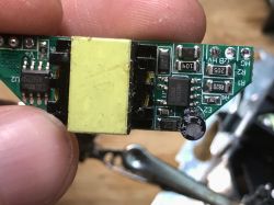 Close-up of a PCB with a small transformer, IC, and electrolytic capacitor, held between fingers.