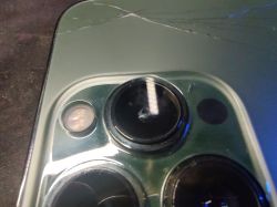Widok zbliżony na uszkodzony aparat iPhone 13 Pro Alpine Green z rysą na obudowie i dziurą w obiektywie.