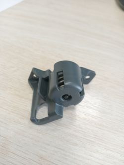 Plastikowy element z ciemnoszarego tworzywa leży na jasnym drewnianym blacie.