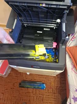Otwarta drukarka Kyocera FS-C2126MFP+ z wyjętym pojemnikiem na toner, wokół widoczny rozsypany toner w kolorze żółtym i niebieskim.