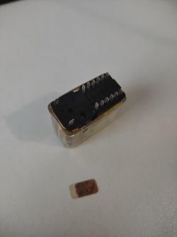 Przekaźnik z czarną podstawą i widocznymi metalowymi pinami leży na białej powierzchni, obok znajduje się mała etykieta.