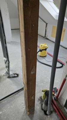 Drewniany słup konstrukcyjny w pomieszczeniu w trakcie remontu, z rurami instalacyjnymi i żółtym odkurzaczem w tle.