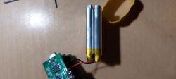 Dwa cylindryczne ogniwa akumulatora połączone równolegle żółtą taśmą, z przylutowanymi przewodami, obok fragment płytki elektronicznej i żółta osłona.