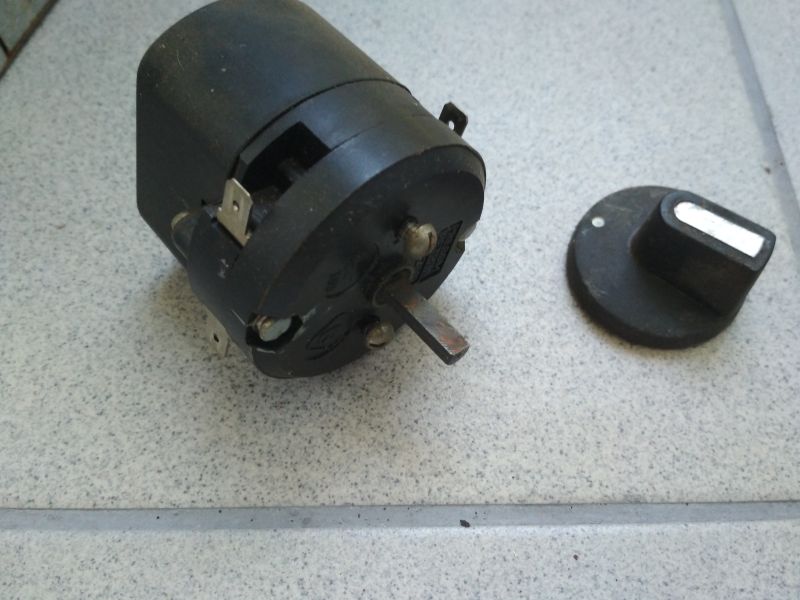 Czarny mechaniczny wyłącznik czasowy oraz gałka z opiekacza Predom Selfa leżące na jasnej powierzchni.